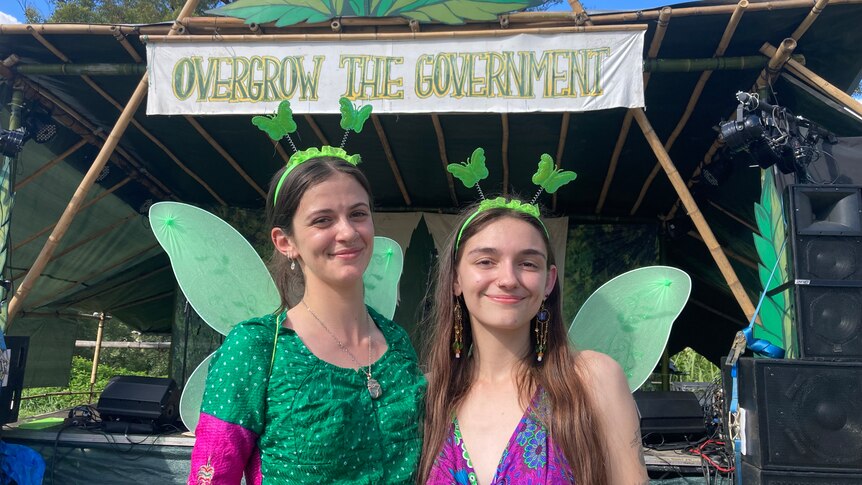 Two girls with green wings on stand in front of a sign reading Overgrow the Government.