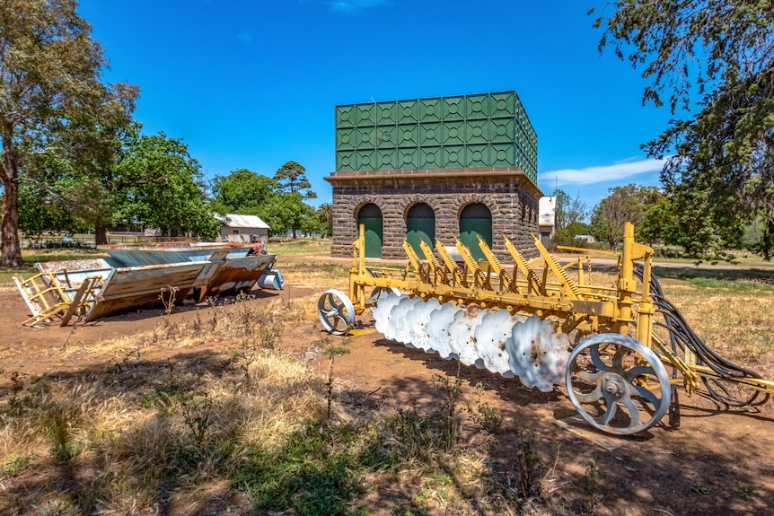 Old rusted farm equipment and a bluestone water tower building surrounded by trees and grass.