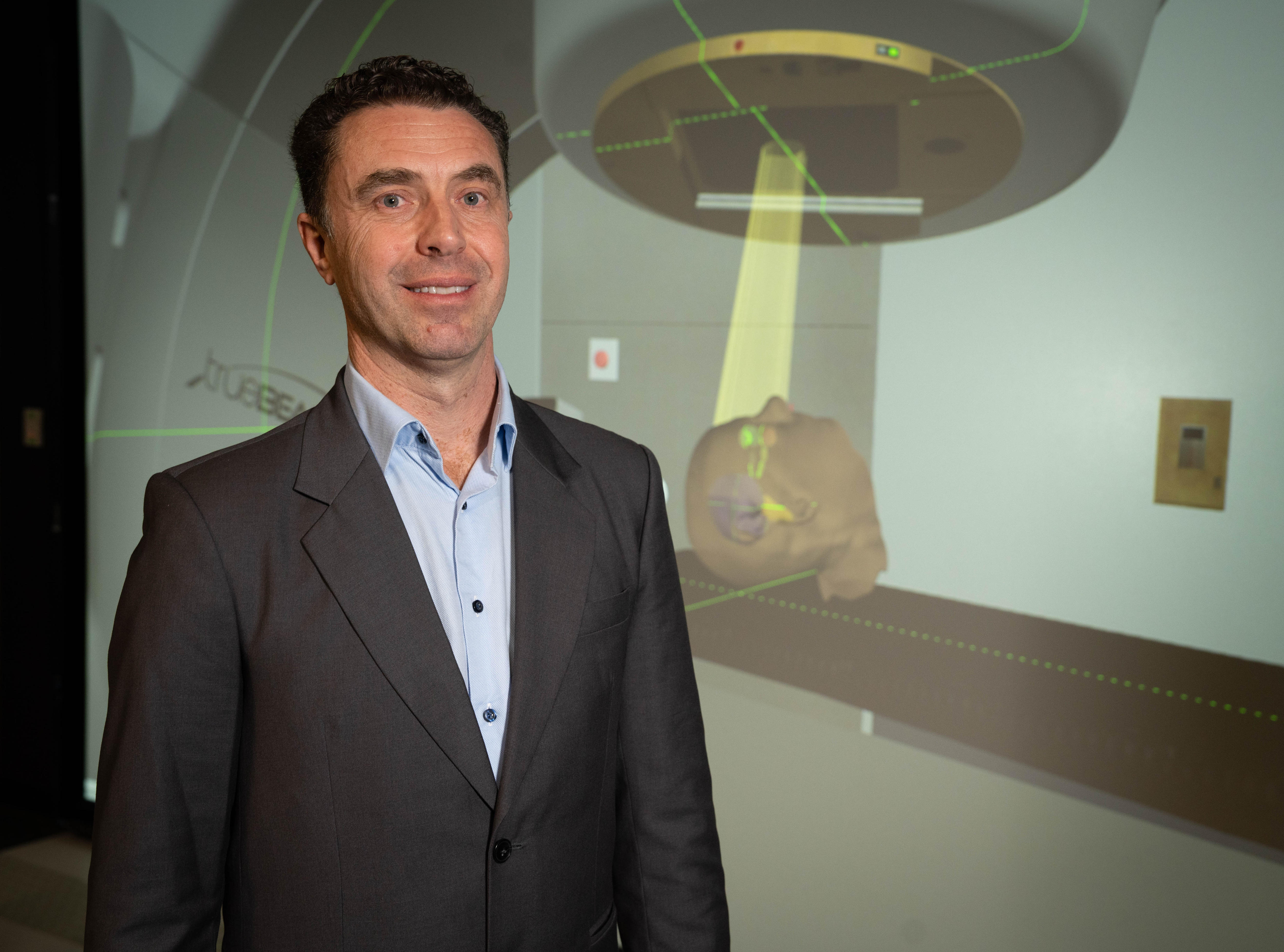 A man wearing a grey suit and blue shirt stands in front of digital projection
