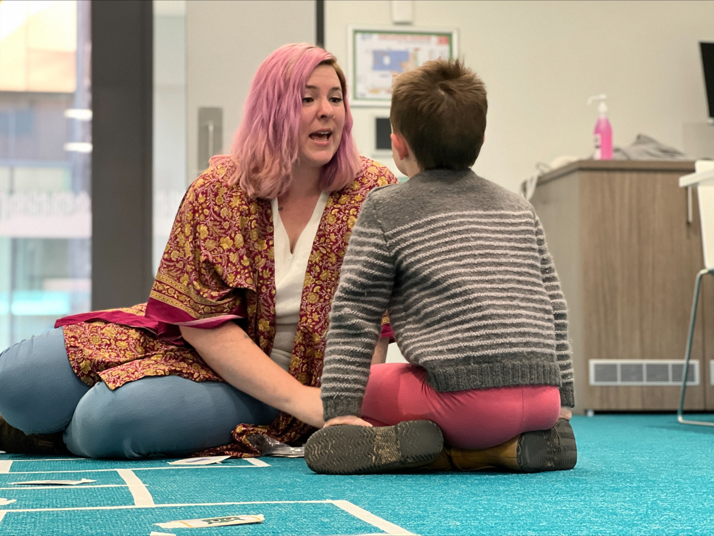 A woman with pink hair and an open mouth sits on the floor facing a boy.