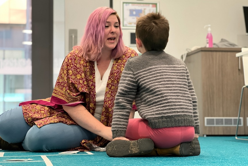 A woman with pink hair and an open mouth sits on the floor facing a boy.