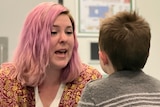 A woman with pink hair and an open mouth sits on the floor facing a boy.