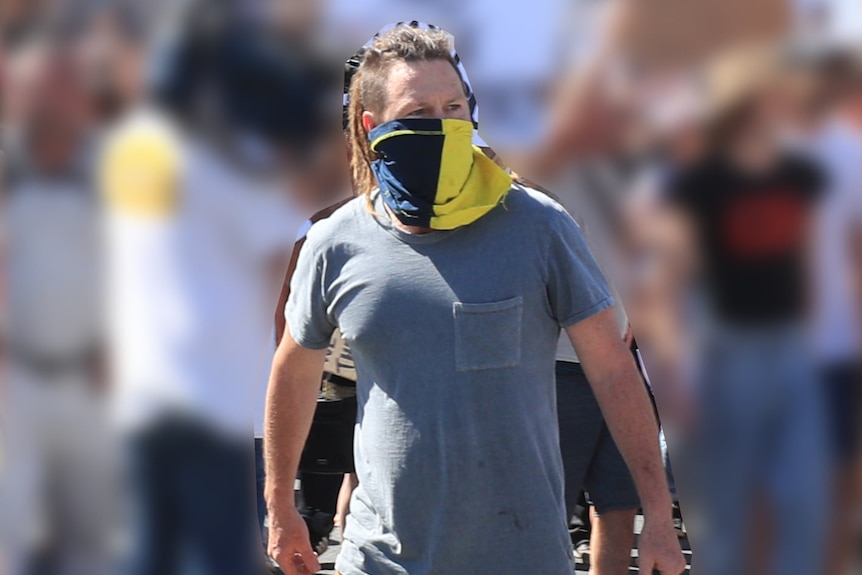 man wearing a faded grey t-shirt, orange pants and a blue and yellow face mask, holding a mobile phone.