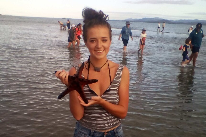 A girl in singlet and shorts stands in knee deep water holding a starfish