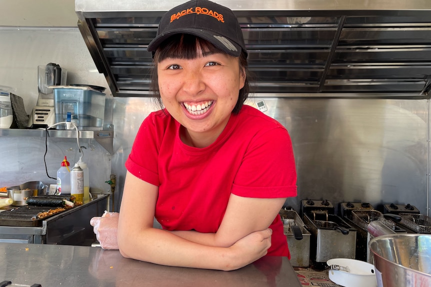 A Taiwanese woman wearing a red shirt and black cap with the words Back Roads emblazoned in orange stands smiling in a food van