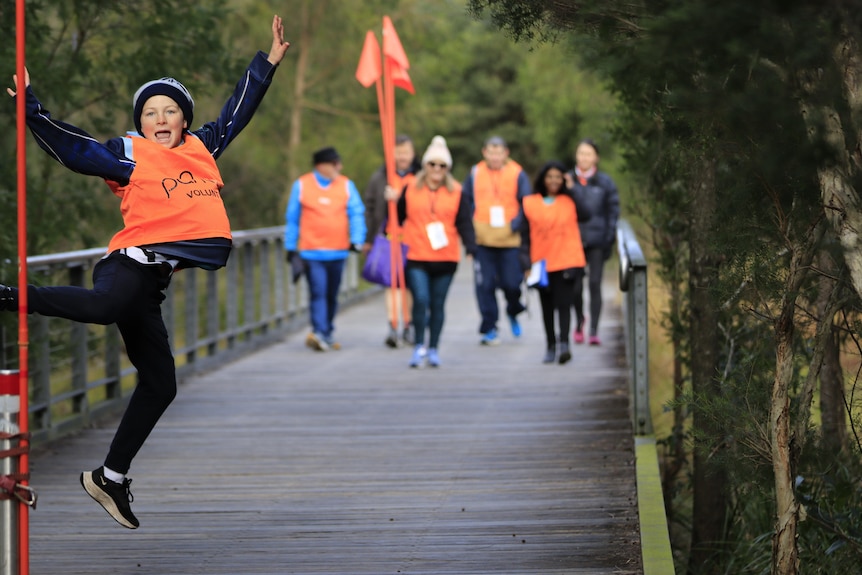 A child in an orange vest jumps as a group of people in the distance walk behind him. 