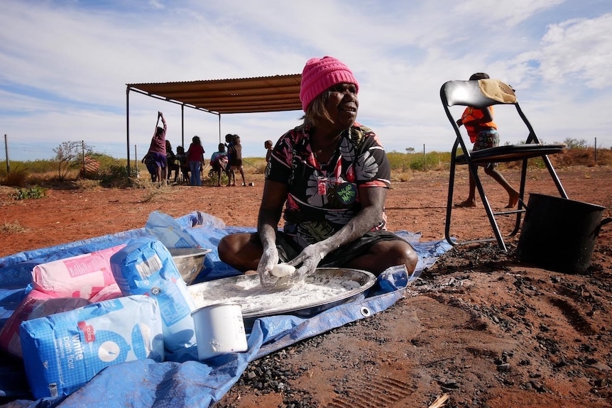 A woman sits on a blue tarp making damper in Balgo, August 2022. 
