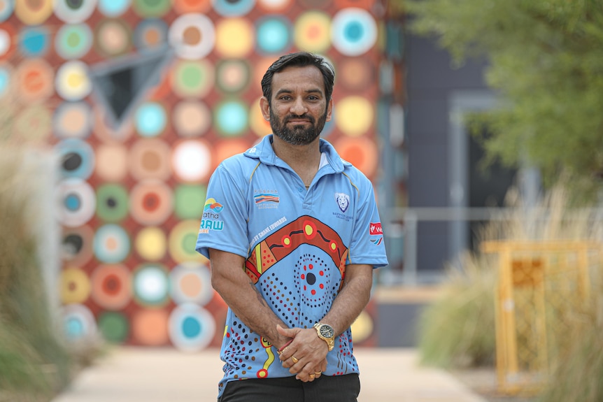 A man in a bright blue cricket shirt smiles at the camera. He stands in front of a patterned wall.