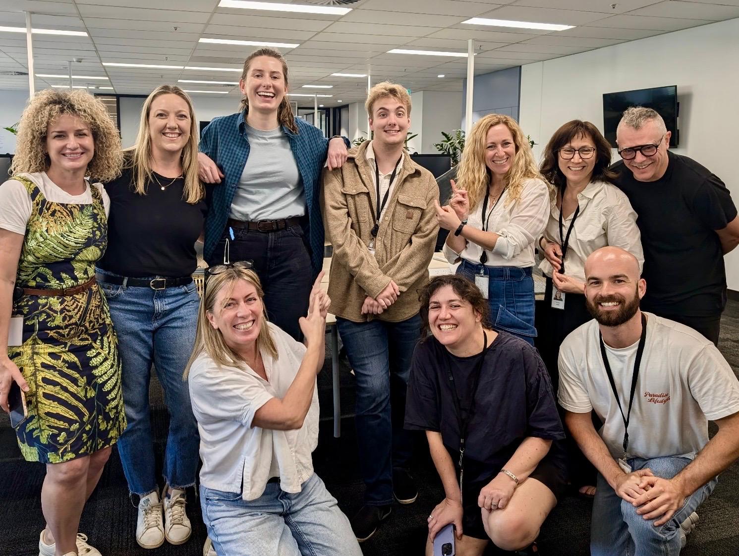 10 cheerful people standing and kneeling in an office, pointing to a to a blond teenage boy in the middle of the group.