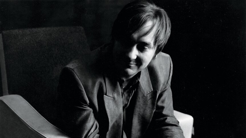 Black and white photo of a smiling Ed Kuepper sitting on a chair