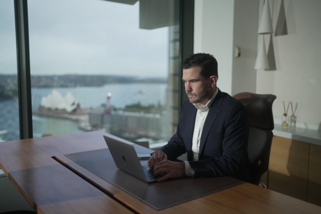 Man sits at desk working on computer in a luxury office. Sydney Opera House and the harbour are visible in the background