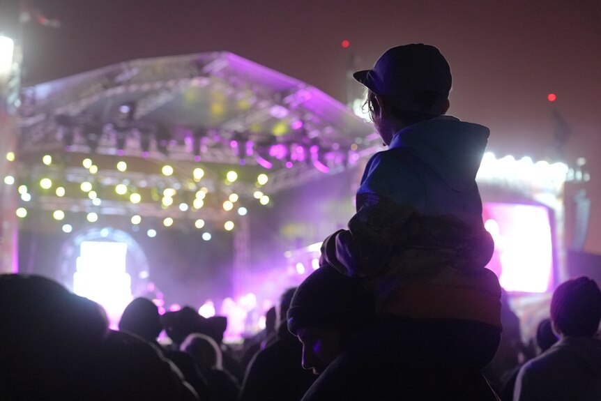 A child on their parent's shoulders views a music festival stage.