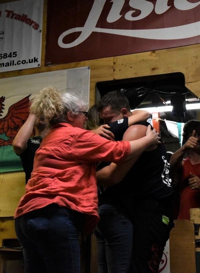 A young man is being hugged by his family in front of a Welsh flag