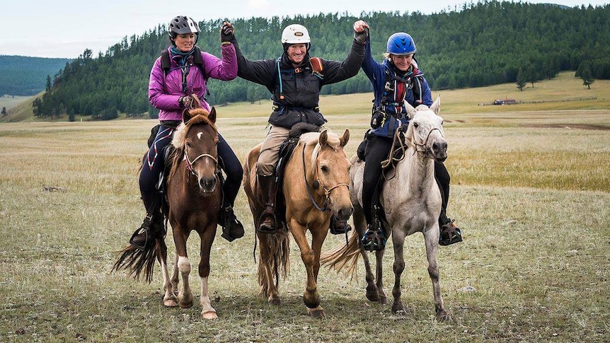 Three tired riders astride small Mongolian ponies with hands upraised in victory