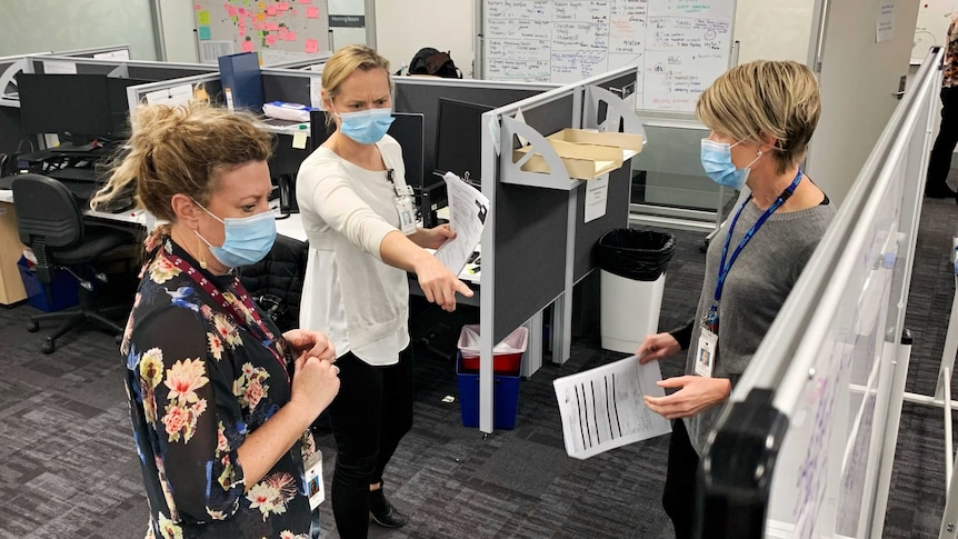 Three women in an office, looking at a whiteboard, one woman points at it, all wearing face masks.
