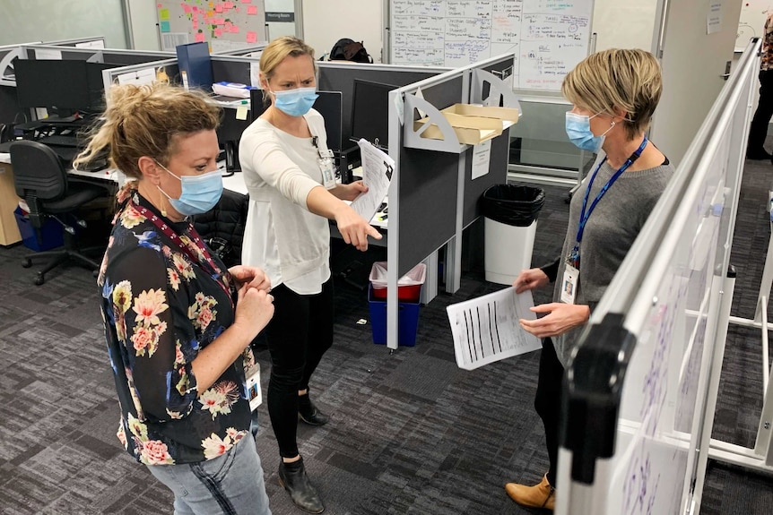 Three women in an office, looking at a whiteboard, one woman points at it, all wearing face masks.