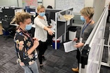 Three women in an office, looking at a whiteboard, one woman points at it, all wearing face masks.