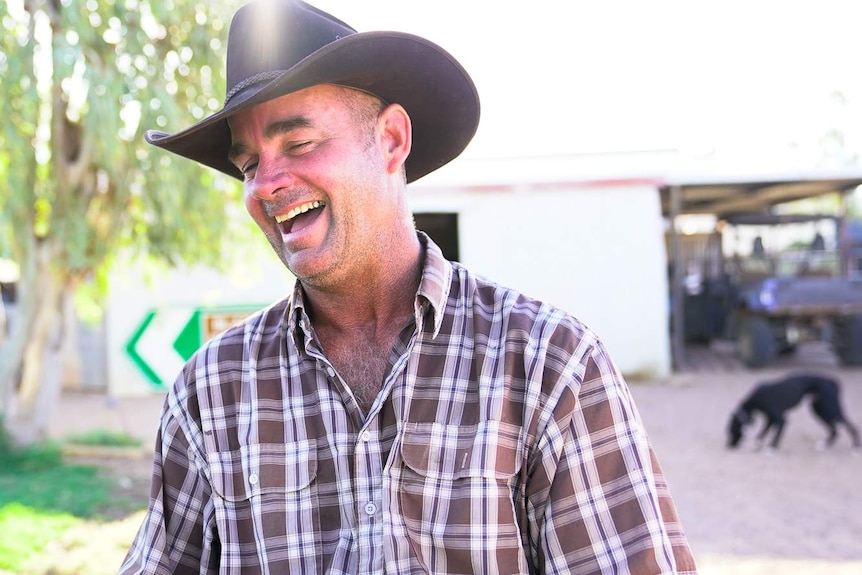 A man in a checked shirt and akubra laughs in the yard of a large outback shed, with dogs running around in the background.