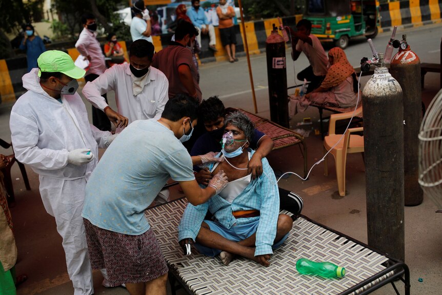A man is helped by health workers as he breaths oxygen through a line connected to a large gas cylinder.