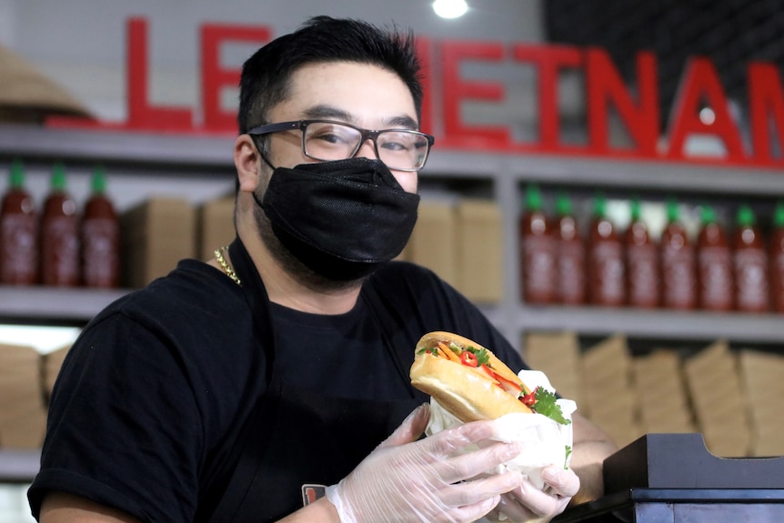 A man wears a black shirt and a facemask while wearing white plastic gloves holding a salad roll. 