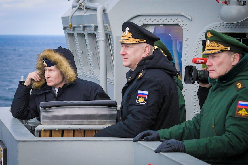 Vladimir Putin stands on a naval ship in a fur trimmed jacket with two military men