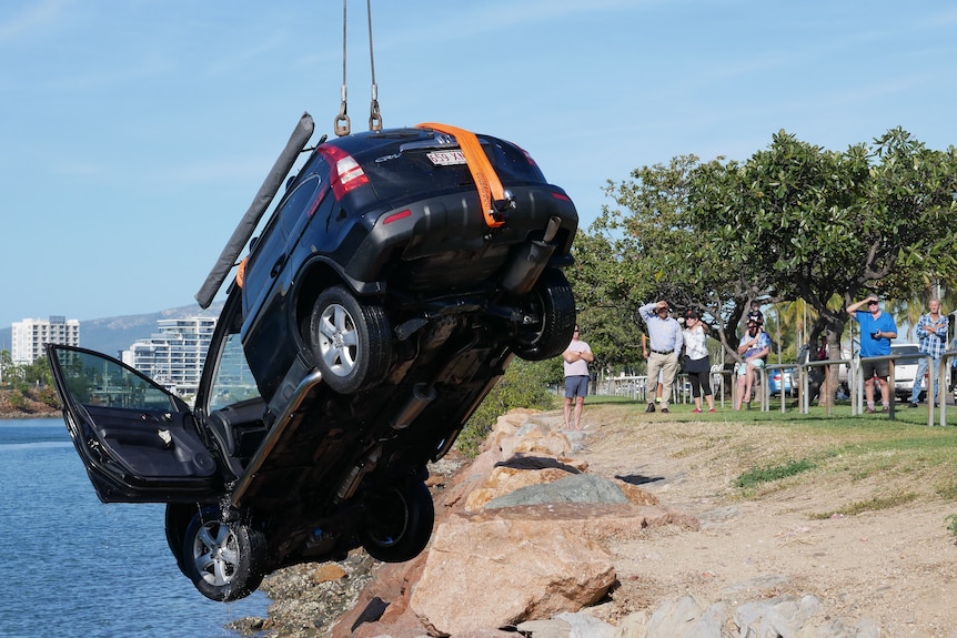 A dark S-U-V being pulled out of a river by a crane as residents watch on