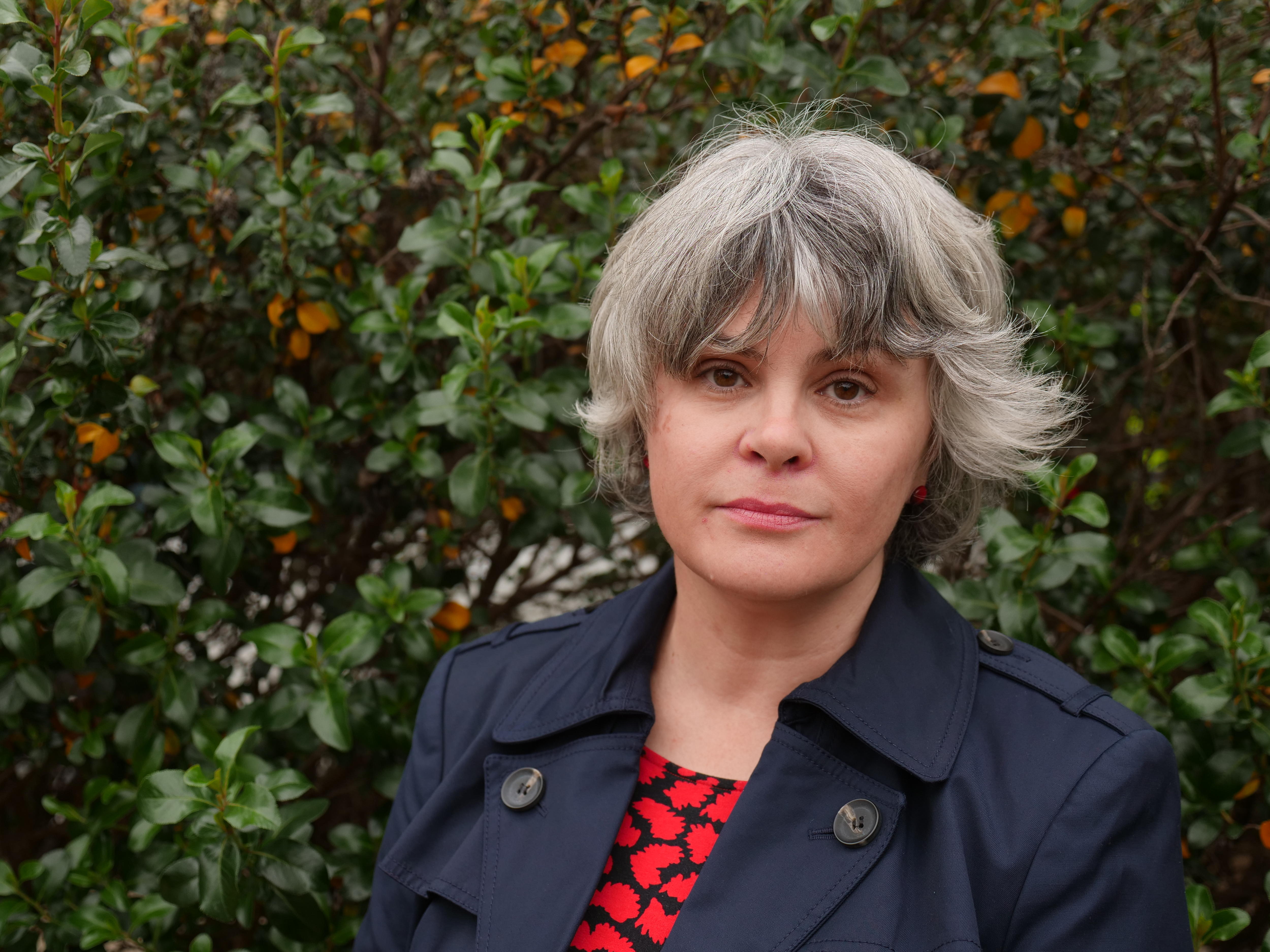 A woman with grey hair looks into camera, there are leaves in the background. 
