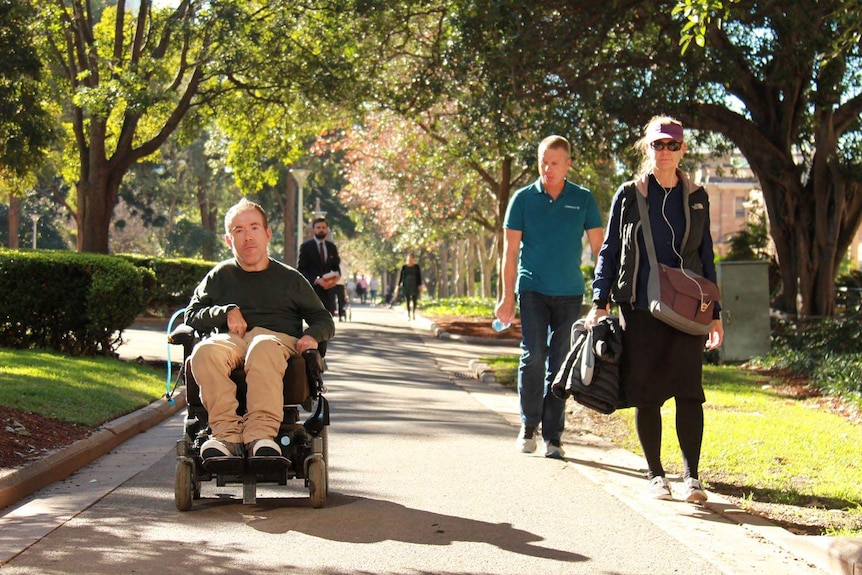 Oliver Morton-Evans pushing his wheelchair along a path in a park, watched by passers-by