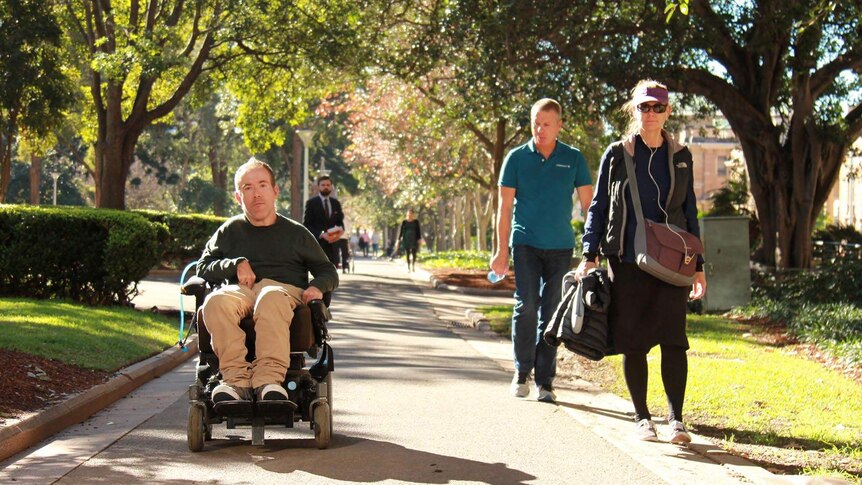 Oliver Morton-Evans pushing his wheelchair along a path in a park, watched by passers-by