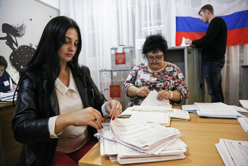 Two seated women and a man standing behind them count votes by hand inside a small office with a Russian flag on the wall. 