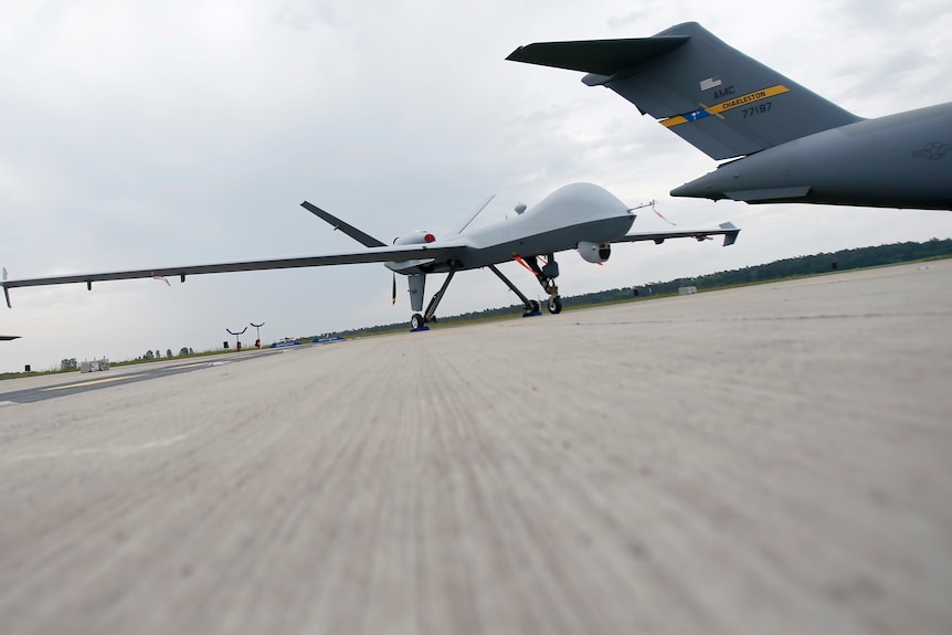 A hi-tech-looking white-grey pilotless drone is parked on an airport tarmac, the picture taken at an angle.