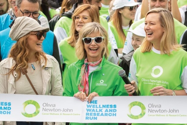 Olivia Newton-John smiling broadly while holding a "Wellness Walk and Research Banner" with a team of walkers.