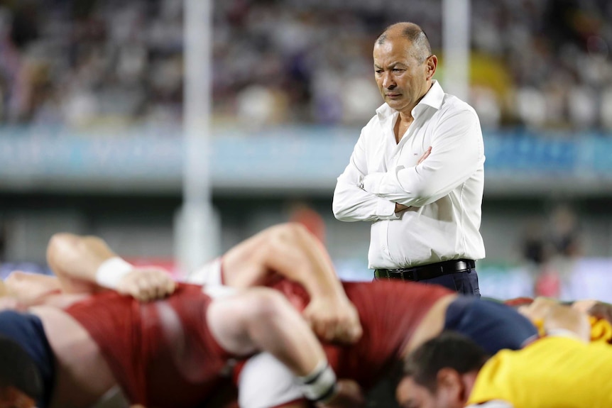 A male rugby union coach stands with his arms folded as he watches his team practise a scrum.