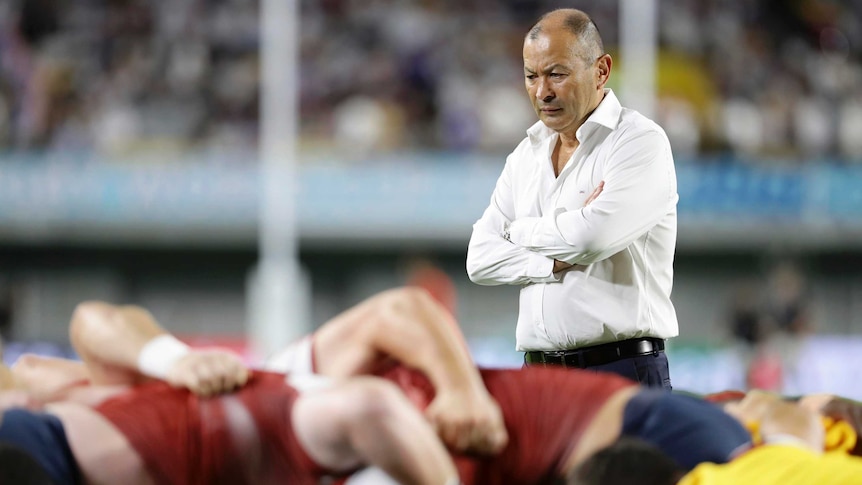 A male rugby union coach stands with his arms folded as he watches his team practise a scrum.