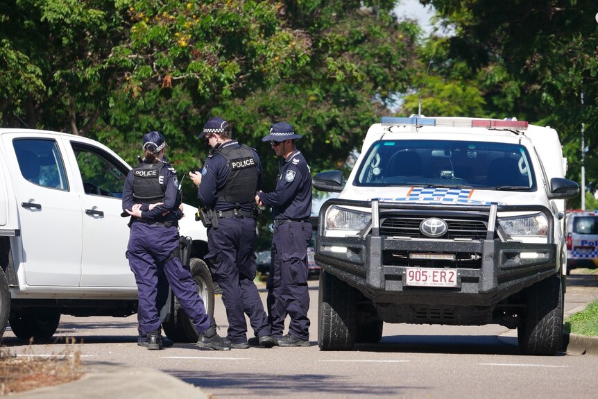 three police officers standing in a group talking between a marked police car and another white vehicle