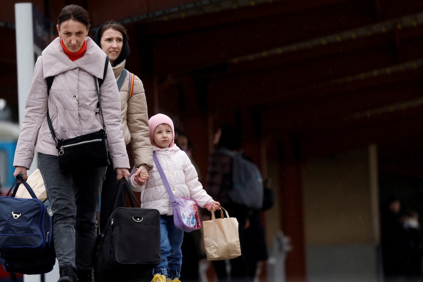 Two women and a girl walk along a train station platform carrying bags.
