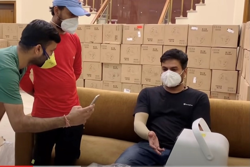 A man in a mask sits on a couch in a room stacked with boxes. 