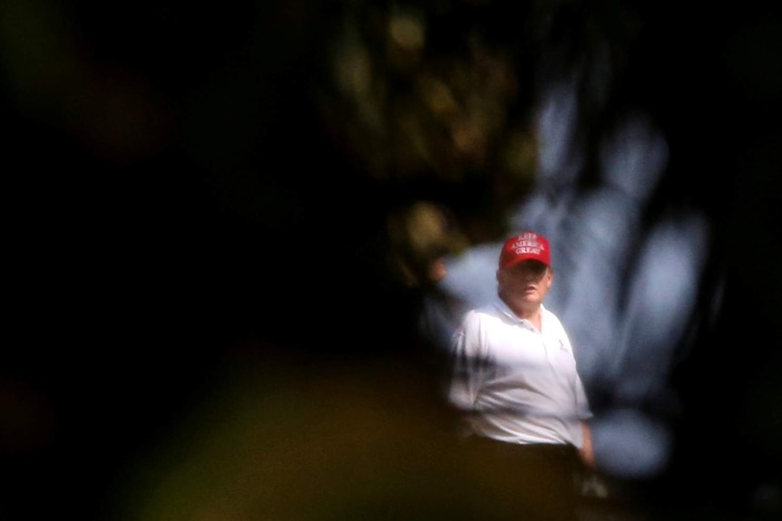 Donald Trump, wearing a white polo shirt and a red Keep America Great cap is framed by trees.