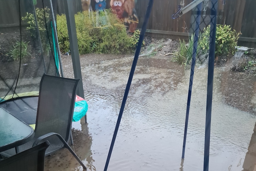 Brown water flows into a backyard through gaps under a wooden fence, pooling around children's toys and outdoor furniture.