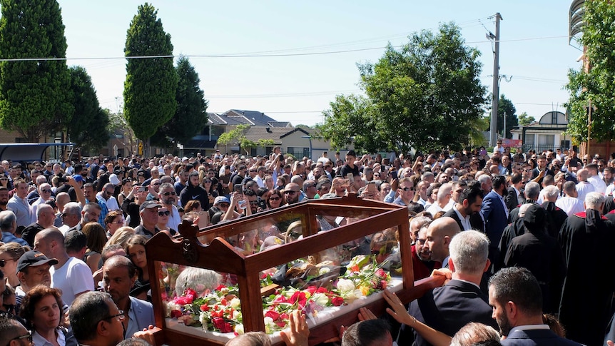 A large crowd of Maronite Catholics watching men carry a glass coffin with flowers inside.