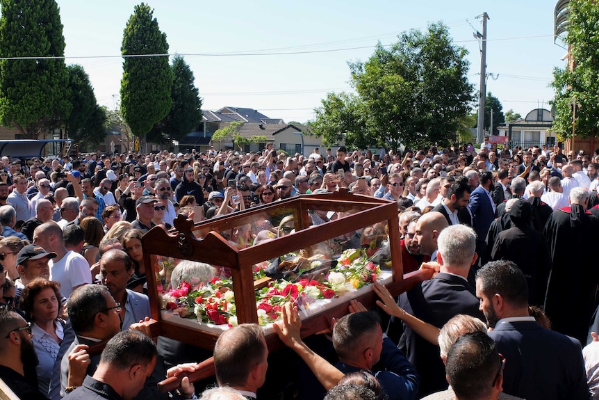 A large crowd of Maronite Catholics watching men carry a glass coffin with flowers inside.
