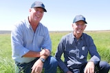Two men kneel in a paddock and smile at the camera