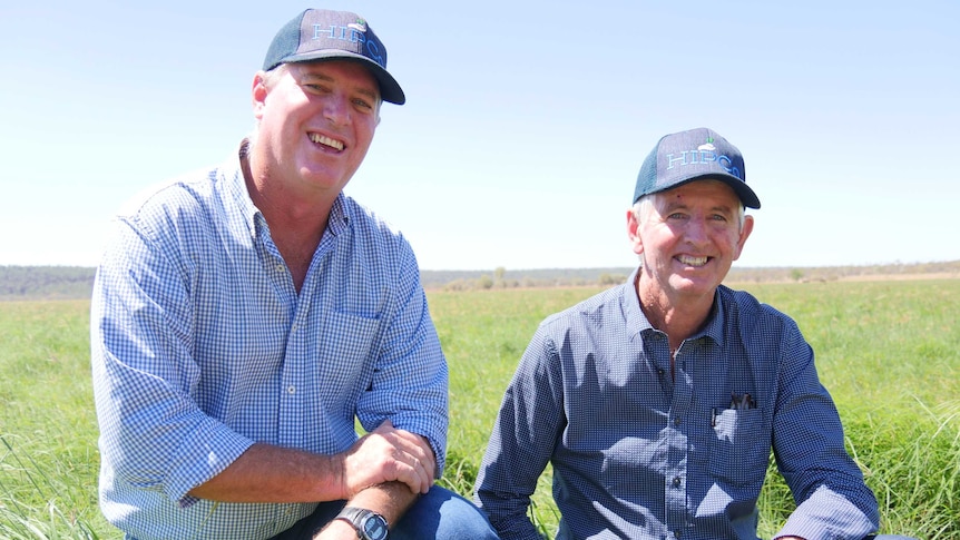 Two men kneel in a paddock and smile at the camera