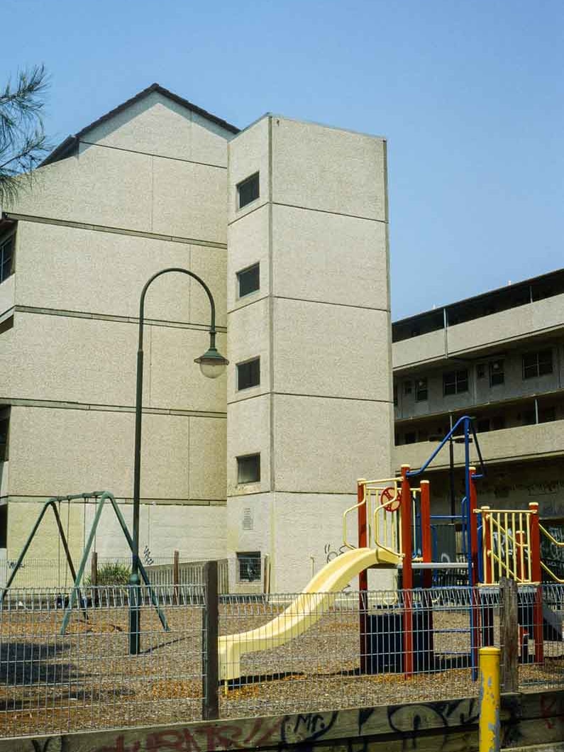 On a clear blue day, you view a 1960s-era public housing estate, made up of low-rise duplexes sitting behind a playground.