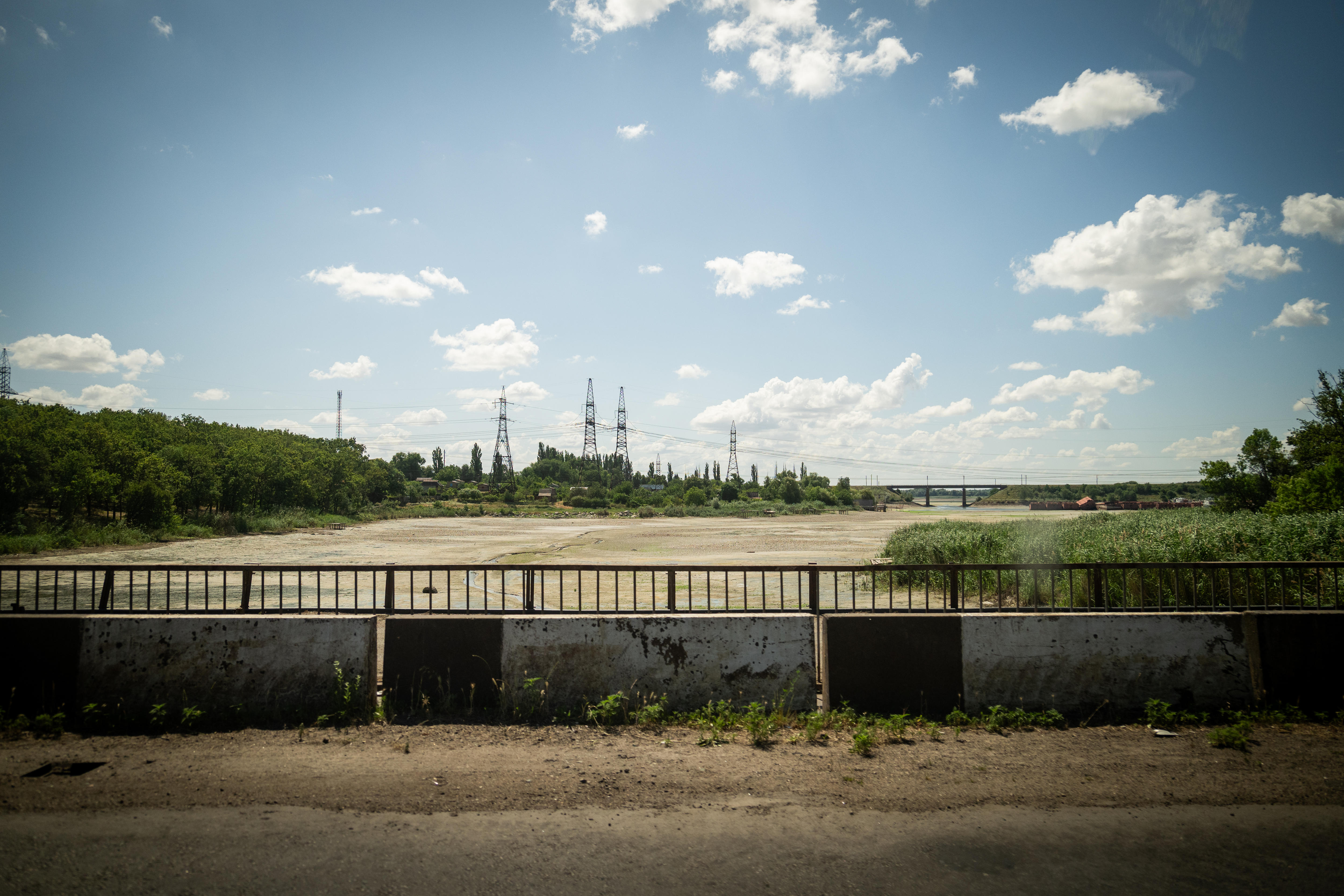 A dry river bed is seen from a dusty bridge. Electricity poles dot the horizon in the distance