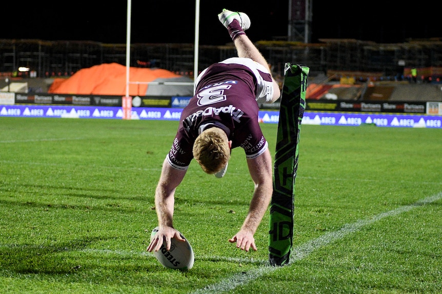 A Manly NRL player dives in the air as he grounds the ball with his right hand to score a try.