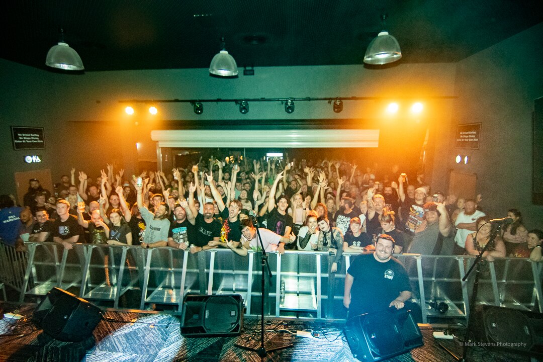 A crowd of people cheer with their arms in the air from behind a crowd barrier inside a small venue.