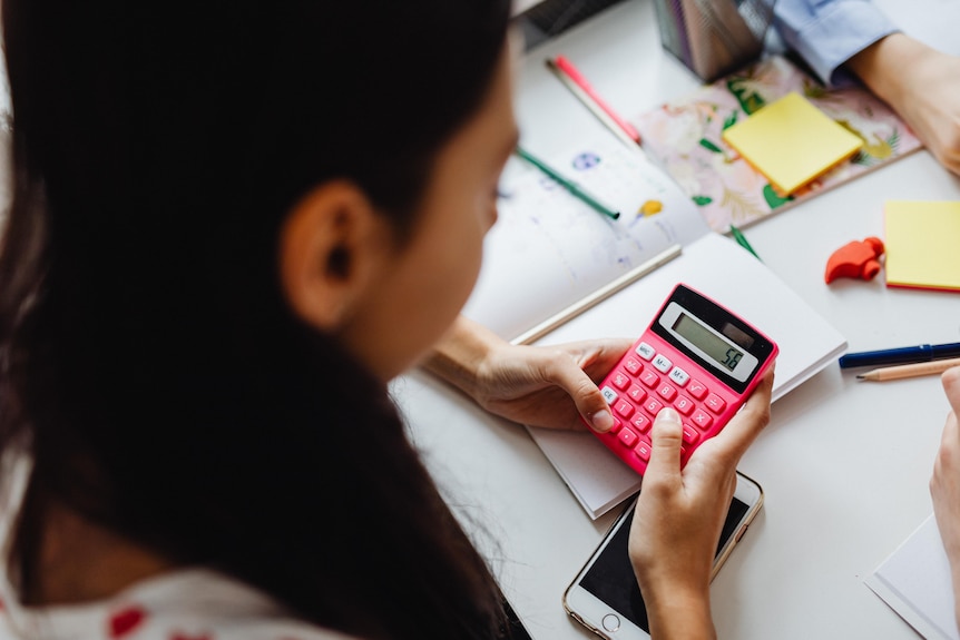 A person with long brown hair uses a calculator at a messy desk. 