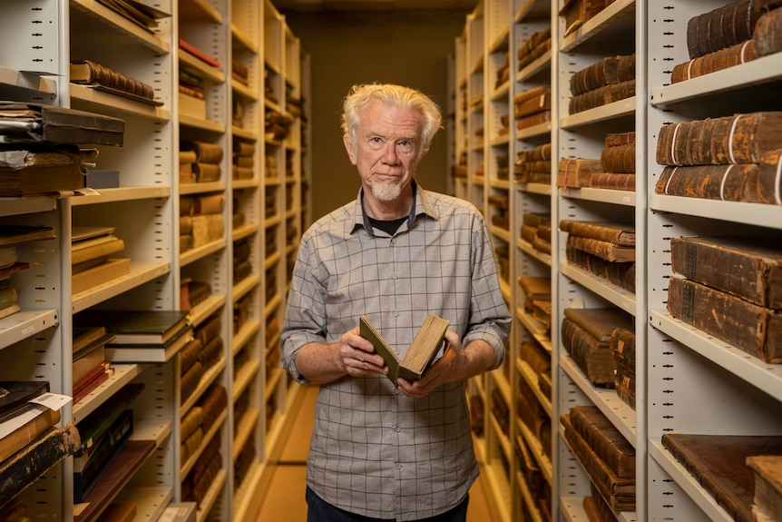 Older man with white hair holds book and looks into camera lens while standing between two shelves loaded with rare books.