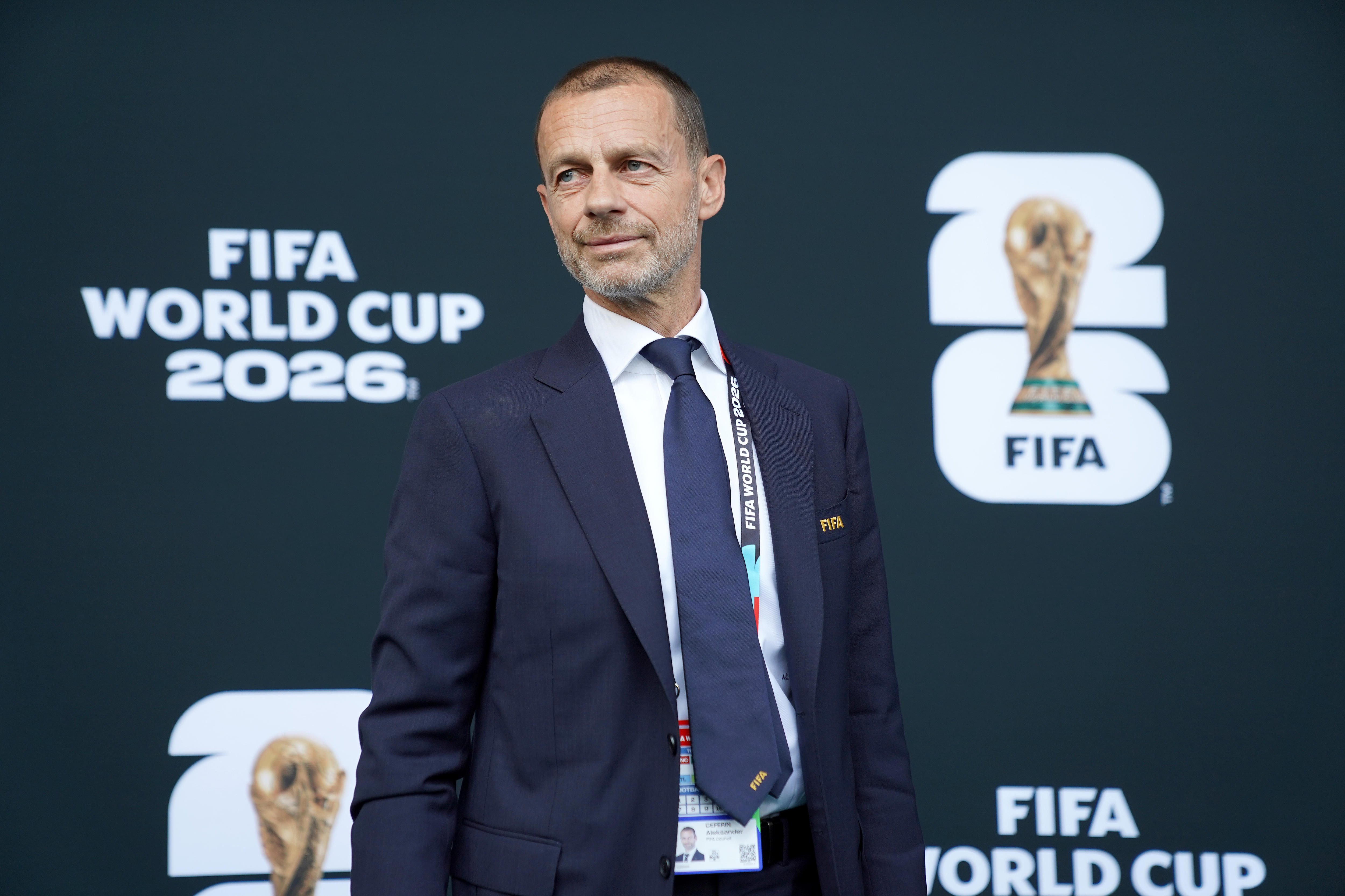Aleksander sorri, olha de lado, usa terno azul, gravata, Copa do Mundo FIFA 2026 atrás, é caucasiano.